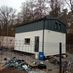 Nowoczesny, dwukondygnacyjny budynek z ciemnym, metalowym dachem i jasną elewacją, ogrodzony siatką. Widoczne drzewa w tle i elementy placu budowy.