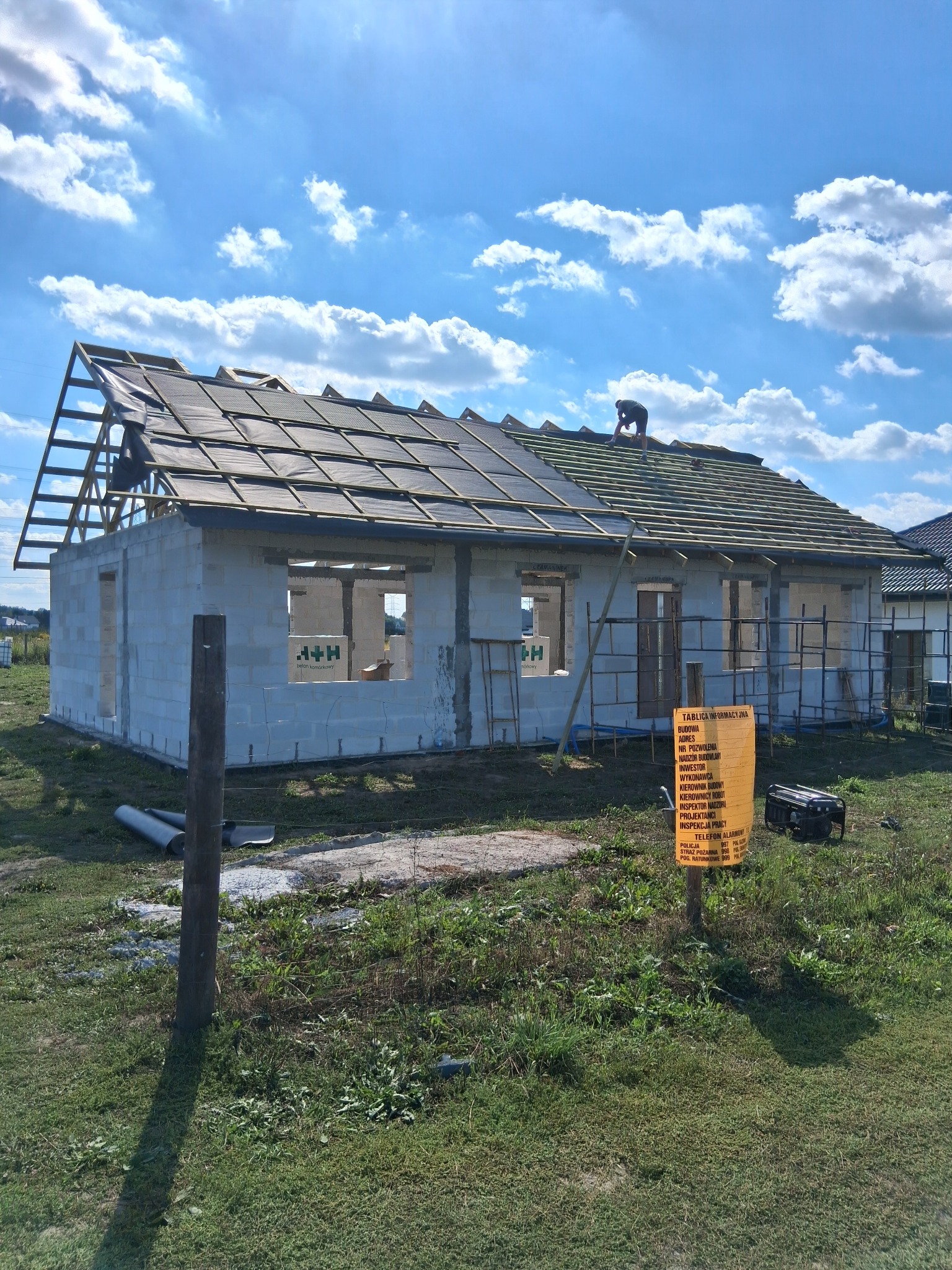 Budowa domu jednorodzinnego w trakcie krycia dachu. Widoczni dekarze na dachu, ściany z betonu komórkowego i tablica informacyjna budowy. Słoneczny dzień.