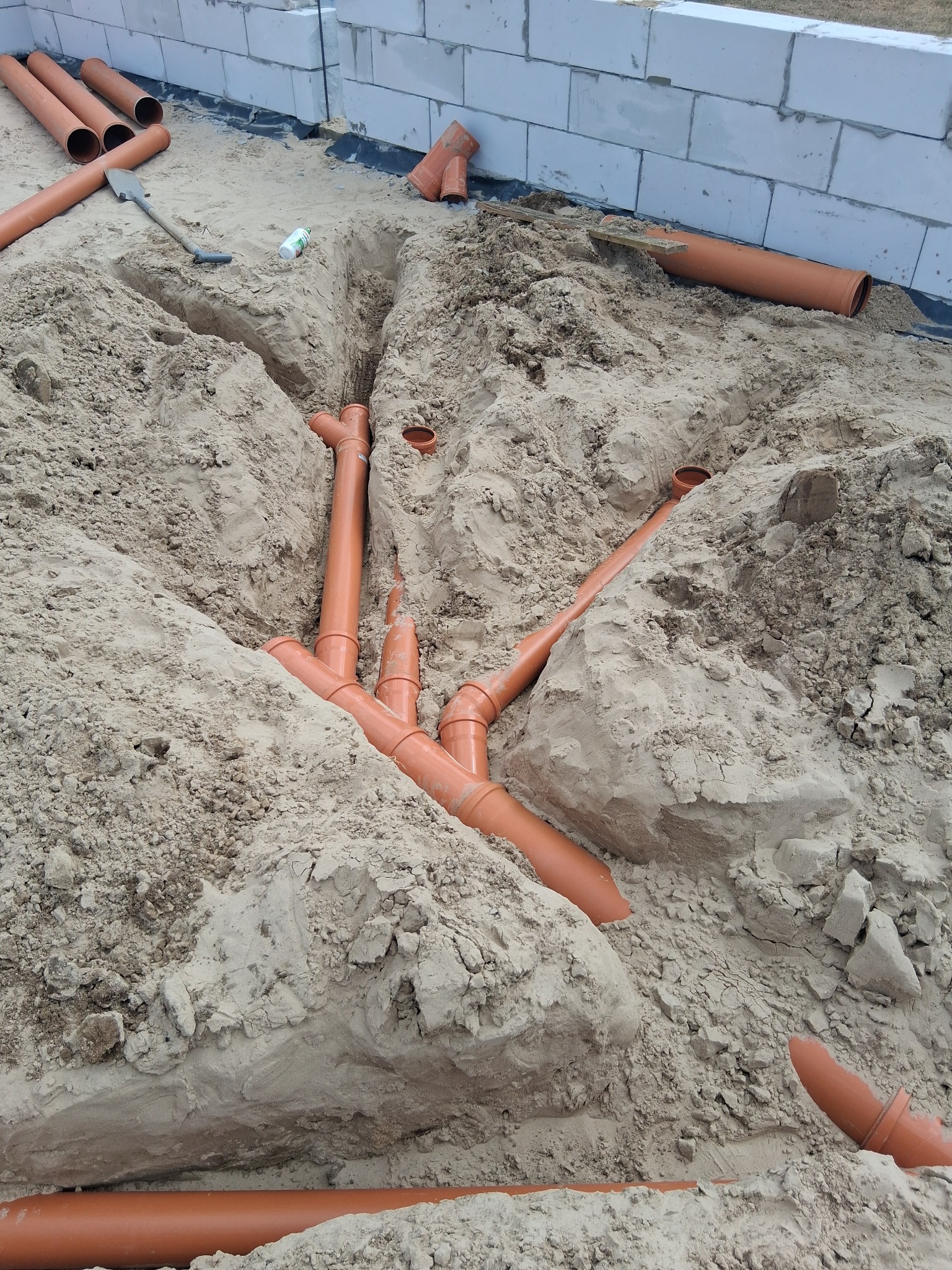 Pomarańczowe rury kanalizacyjne ułożone w wykopie z piasku, widoczne białe bloczki fundamentowe w tle. Etap prac instalacyjnych przed zasypaniem.