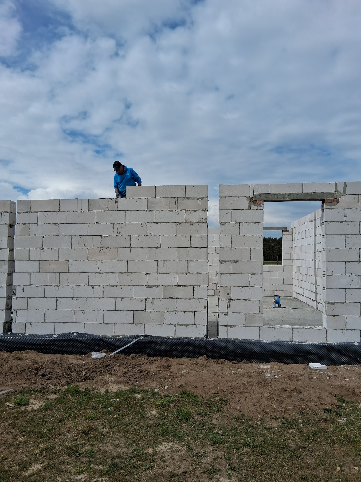 Wznoszenie ścian z białych bloczków betonu komórkowego. Widoczny fragment ściany z otworem okiennym i pracownik budowlany na szczycie muru. Budowa domu w toku.