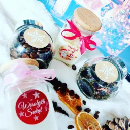 Cztery szklane słoiki z drewnianymi pokrywkami i świątecznymi etykietami, wypełnione kawą, białym kremem, suszem i białym woskiem, ozdobione wstążkami, leżące na białym futrze z plasterkami...