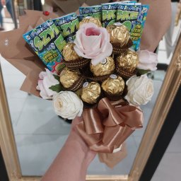 P.H.U. Termix A.J-BUD Jacek Ciekot - Bukiet z róż, czekoladek Ferrero Rocher i losów loterii Szczęśliwy Los, owinięty w brązowy papier, trzymany w dłoni z tatuażem na tle lustra w złotej ramie.