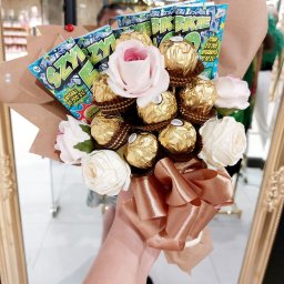 P.H.U. Termix A.J-BUD Jacek Ciekot - Oryginalny bukiet z czekoladek Ferrero Rocher, sztucznych róż i zdrapek lotto Szczęśliwy Los, owinięty w brązowy papier i przewiązany kokardą w kolorze różowego złota, trzymany w dłoni na tle lustra.