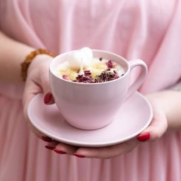 P.H.U. Termix A.J-BUD Jacek Ciekot - Ręce kobiety w różowej sukience trzymają filiżankę z napojem udekorowanym płatkami róż, złotym brokatem i woskowym sercem.