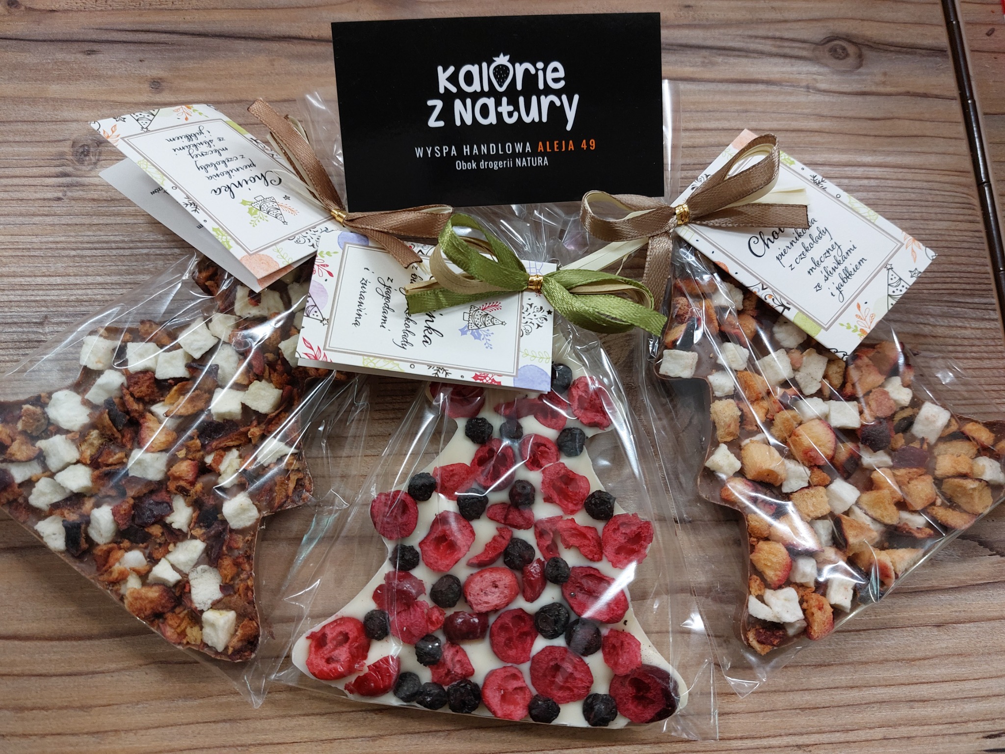 Trzy zapakowane, ręcznie robione czekolady: dwie w kształcie gwiazdy z kawałkami owoców i jedna w kształcie choinki z czerwonymi i czarnymi jagodami, z kartonikiem reklamowym 'Kalorie z Natury'.