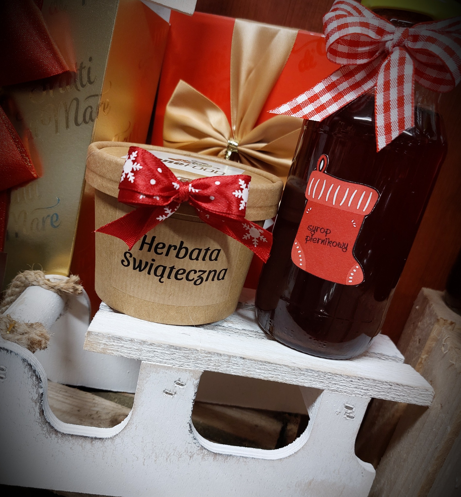 Świąteczny zestaw upominkowy: słoik syropu piernikowego z etykietą w kształcie skarpety i wstążką w kratkę, obok pudełko herbaty świątecznej z czerwoną wstążką w śnieżynki, całość na białych...