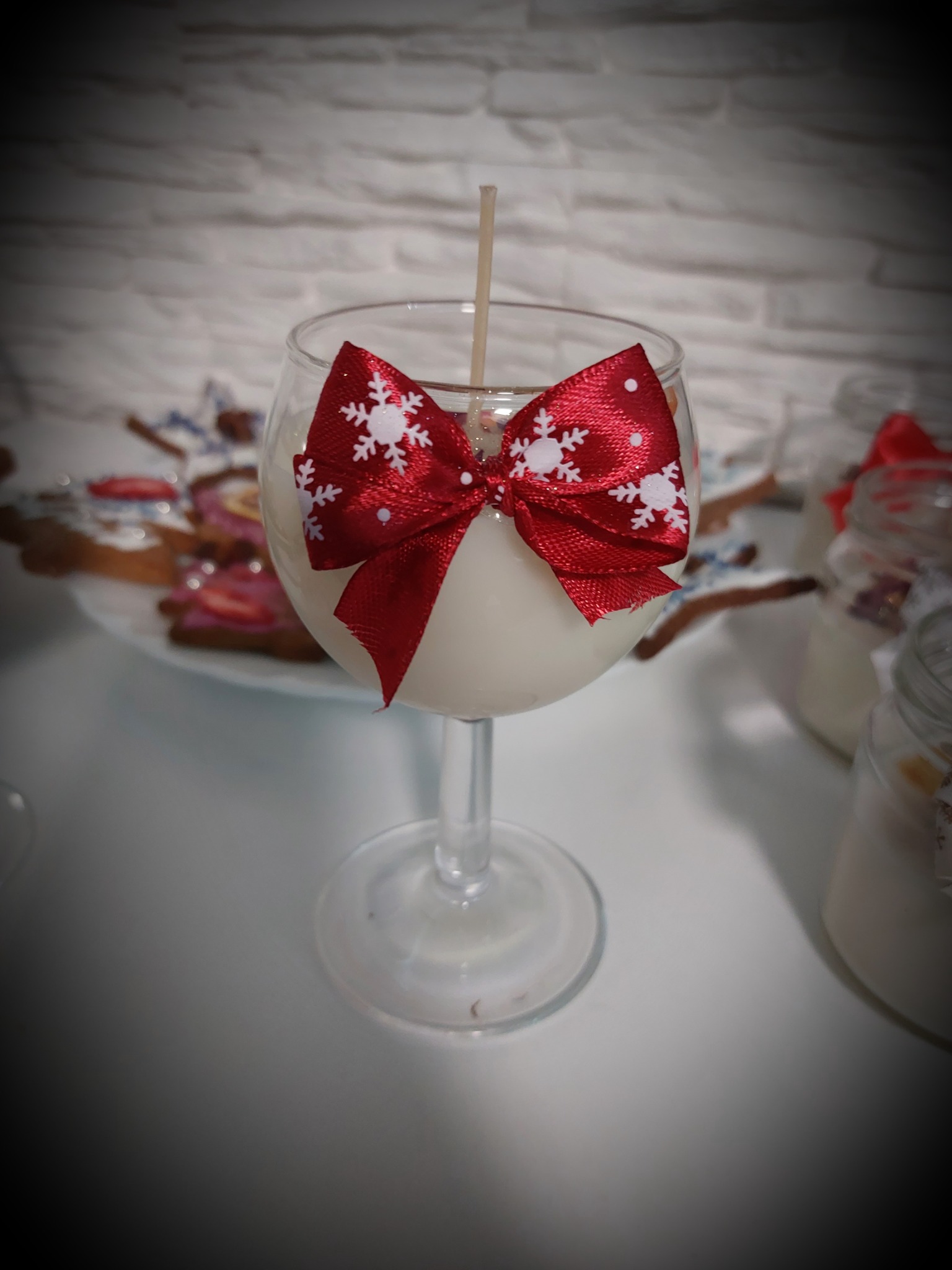 Dekoracyjna świeca w szklanym kieliszku, przewiązana czerwoną kokardą w białe śnieżynki, ustawiona na białym stole z innymi świecami i ciasteczkami w tle.