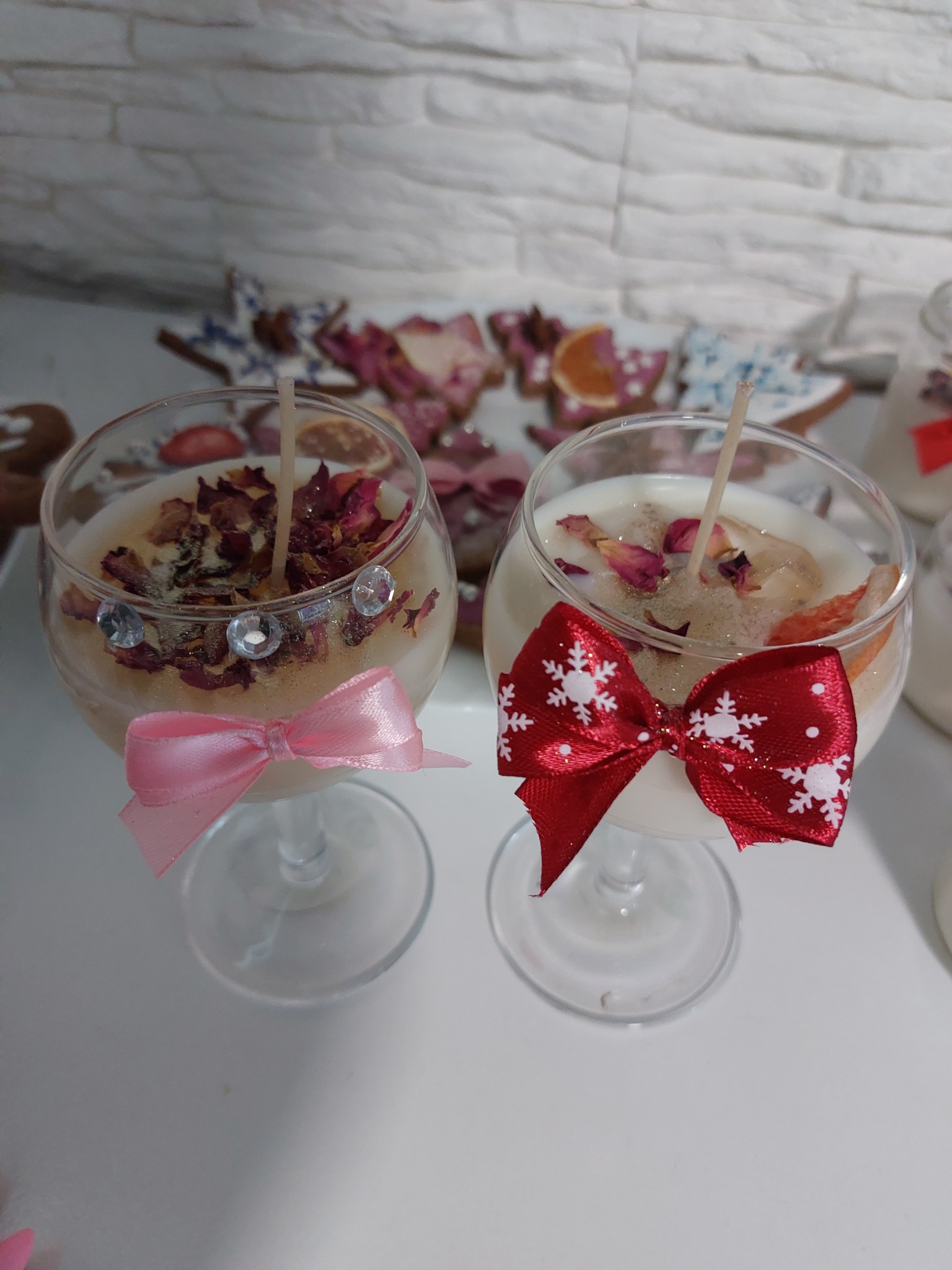 Dwie dekoracyjne świece w szklanych kieliszkach, jedna z różową kokardką i kryształkami, druga z czerwoną kokardką w śnieżynki, posypane płatkami róż i suszonymi owocami.