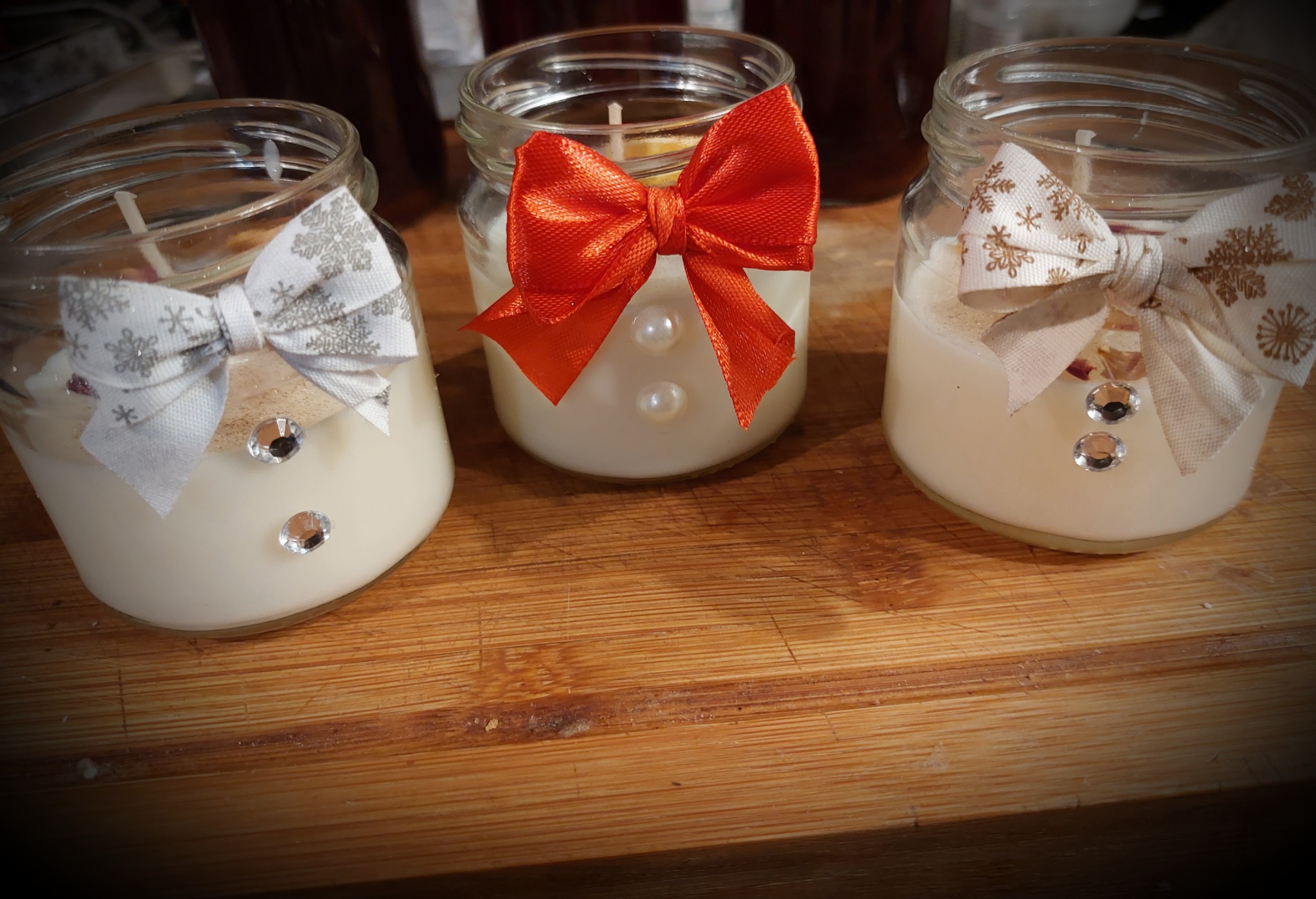 Trzy ozdobne świece w słoikach, każda z kokardą (czerwona i dwie w odcieniach beżu) i dwoma błyszczącymi kamykami, stojące na drewnianym blacie.