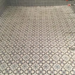 Wykończona podłoga w kabinie prysznicowej z wzorem czterolistnej koniczyny w odcieniach szarości, błękitu i czerwieni, kontrastująca z szarymi, kwadratowymi płytkami ściennymi.