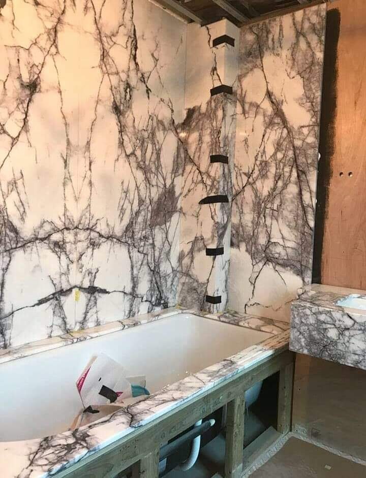 Biała wanna obudowana marmurkową płytą, ściany wykończone tą samą płytą, imitacja marmuru, widoczne czarne taśmy mocujące na narożniku, etap prac wykończeniowych.