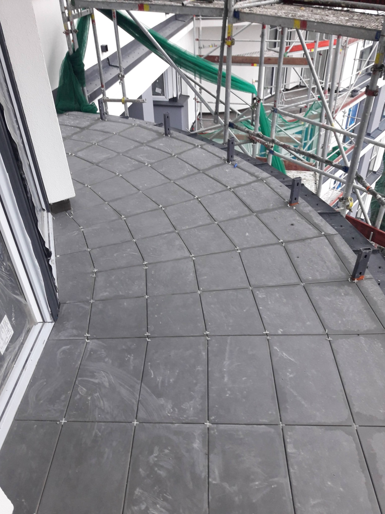 Szare płytki ułożone na balkonie, widoczne krzyżyki dystansowe, rusztowanie w tle.