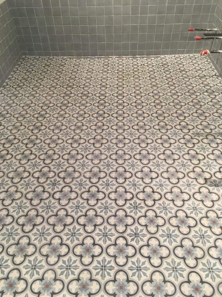 Wykończona podłoga w kabinie prysznicowej z wzorem czterolistnej koniczyny w odcieniach szarości, błękitu i czerwieni, kontrastująca z szarymi, kwadratowymi płytkami ściennymi.