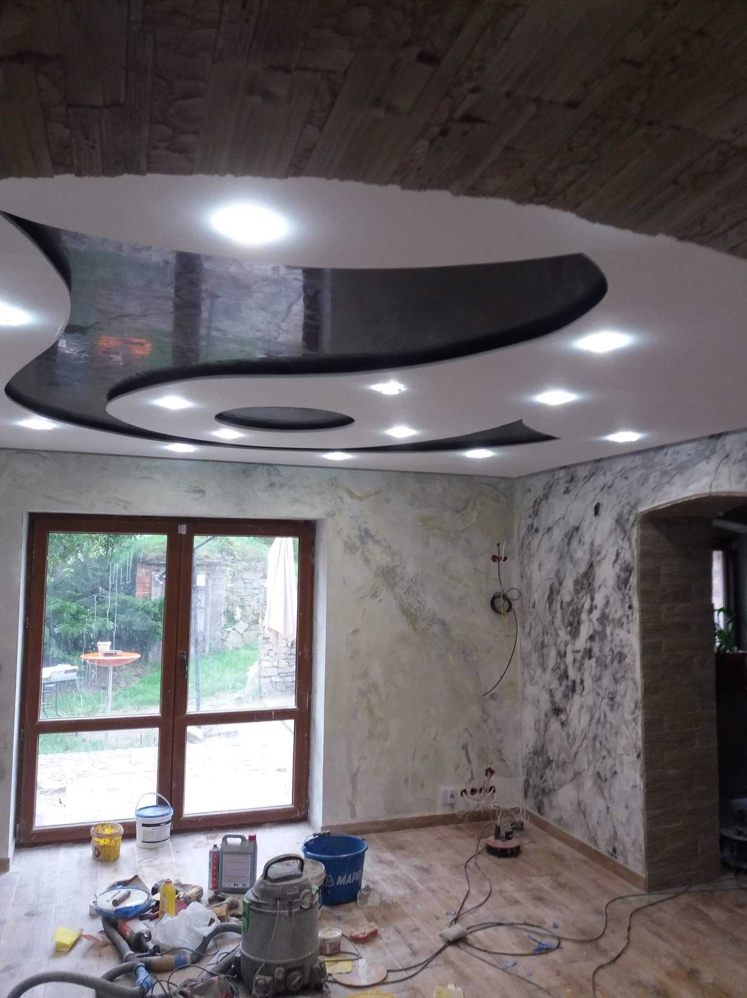 Wnętrze w trakcie remontu: Sufit z gładzią i wbudowanym oświetleniem, ściany z efektem marmuru, widoczne narzędzia i materiały budowlane na podłodze.