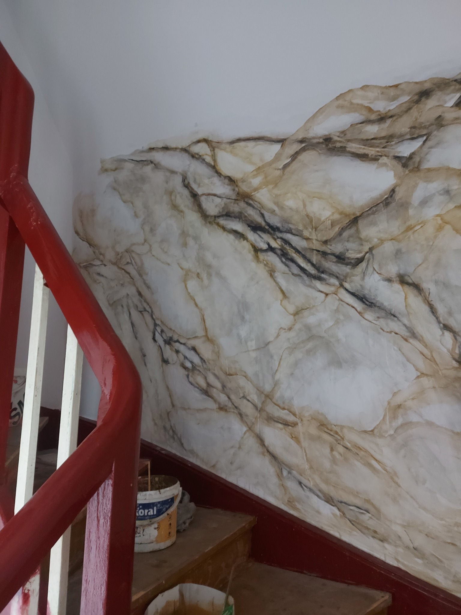 Ściana wewnętrzna z efektem marmuru, wykonana techniką dekoracyjną, widoczna obok czerwonej balustrady schodów, z wiadrami farby u podstawy schodów.