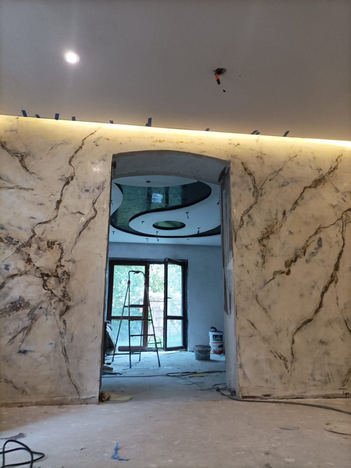 Ściana z efektem marmuru, oświetlenie LED przy suficie, widoczny fragment sufitu z dekoracyjnym wzorem Yin Yang, drabina oraz wiaderka farby w tle, pomieszczenie w trakcie prac wykończeniowych.