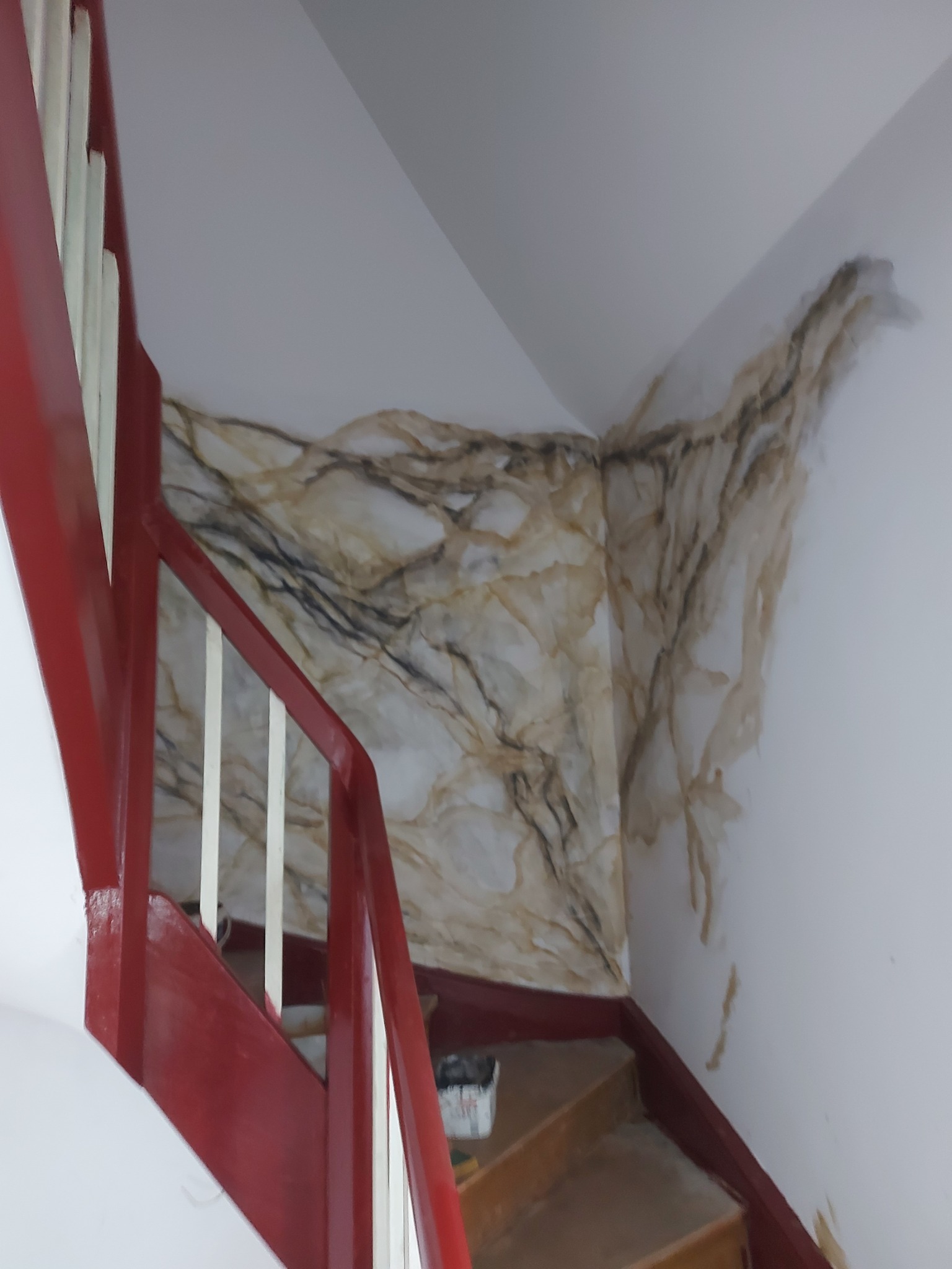 Narożnik ściany przy schodach z widocznymi zaciekami i plamami po zalaniu, imitującymi marmurkowe wzory, czerwona balustrada schodów w kadrze.
