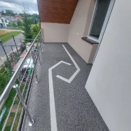 Szara posadzka żywiczna na balkonie z białym geometrycznym wzorem, widok z góry, metalowa balustrada, okno dachowe i drewniana podbitka dachu.