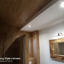 Wykończona łazienka z drewnianym sufitem, kabiną prysznicową z elementami drewna oraz lustrem w ozdobnej drewnianej ramie, oświetlona punktowo i listwą nad lustrem.