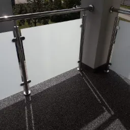 Nowoczesny balkon z podłogą z drobnego kamienia i balustradą ze stali nierdzewnej z matowym szkłem, widok z góry, oświetlony promieniami słonecznymi tworzącymi wzory cieni.