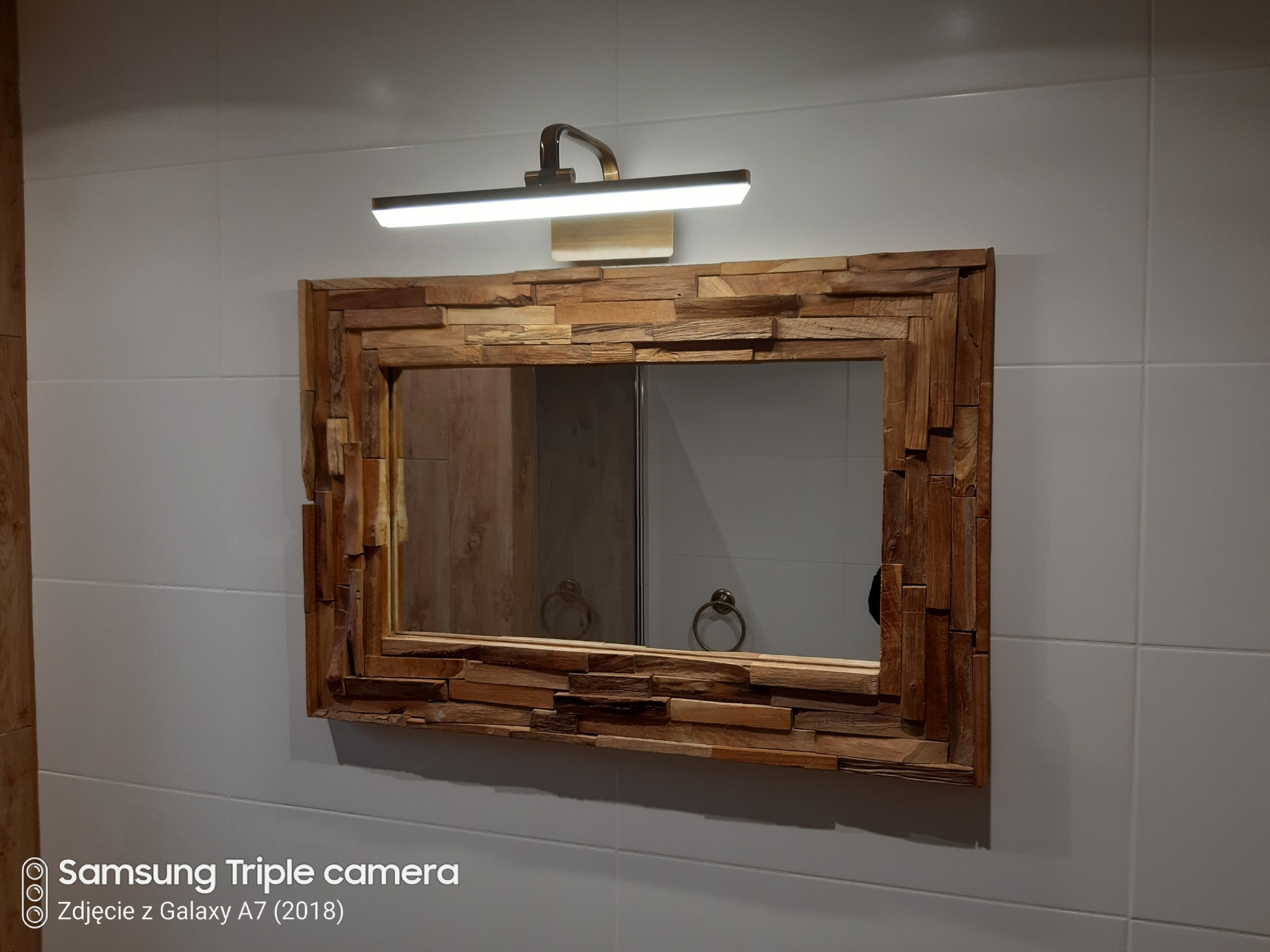 Łazienkowe lustro w ramie z nieregularnych kawałków drewna, oświetlone lampą LED, zawieszone na ścianie z białych płytek.