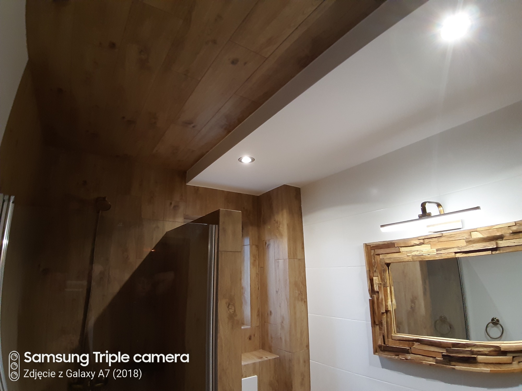 Wykończona łazienka z drewnianym sufitem, kabiną prysznicową z elementami drewna oraz lustrem w ozdobnej drewnianej ramie, oświetlona punktowo i listwą nad lustrem.
