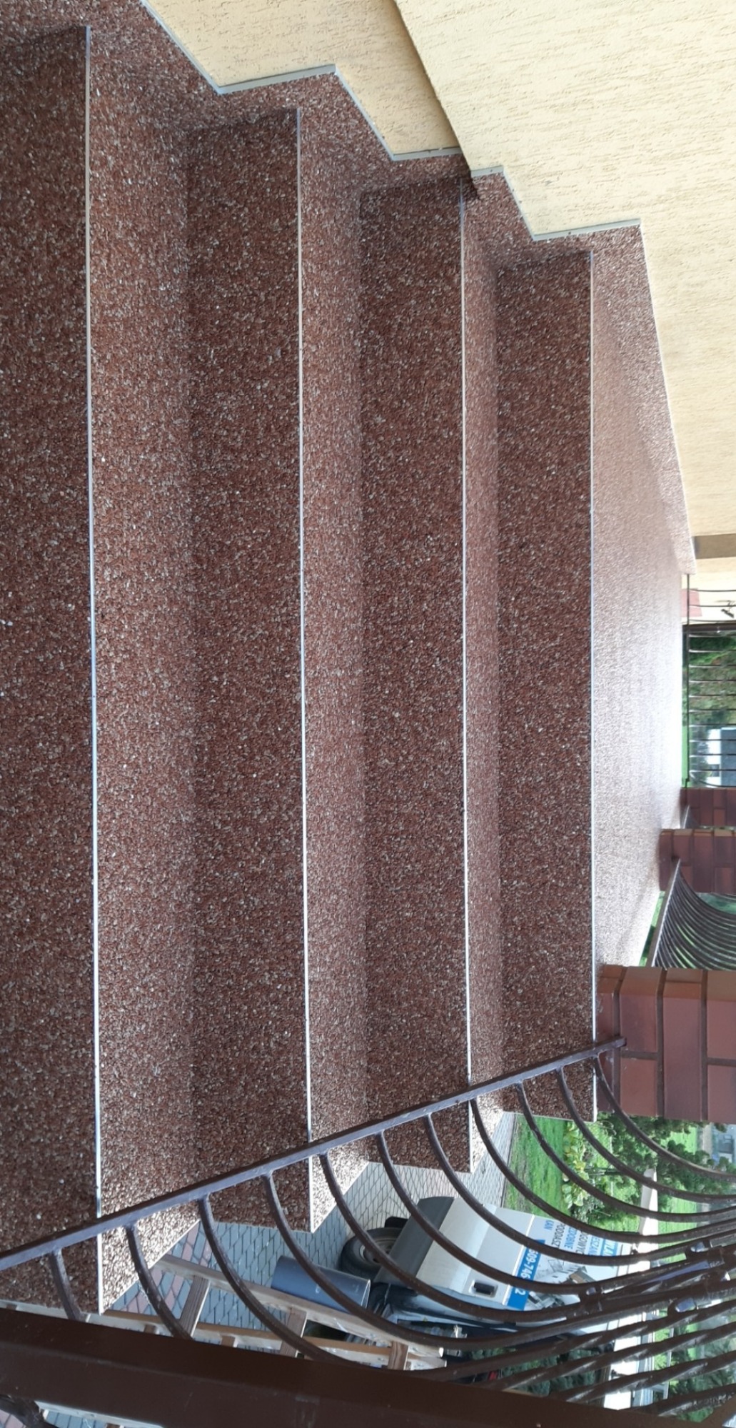 Schody zewnętrzne wykończone brązowym kamyczkowym tynkiem żywicznym, widok z góry na balustradę i zaparkowany samochód w tle.