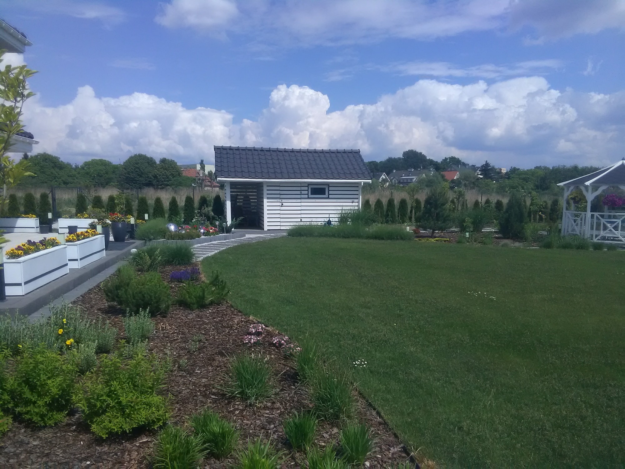 Zadbany ogród z równo przystrzyżonym trawnikiem, ozdobnymi rabatami z korą, białymi donicami z kwiatami, ścieżką z jasnych płyt prowadzącą do białej altany z ciemnym dachem oraz białej, otwartej...