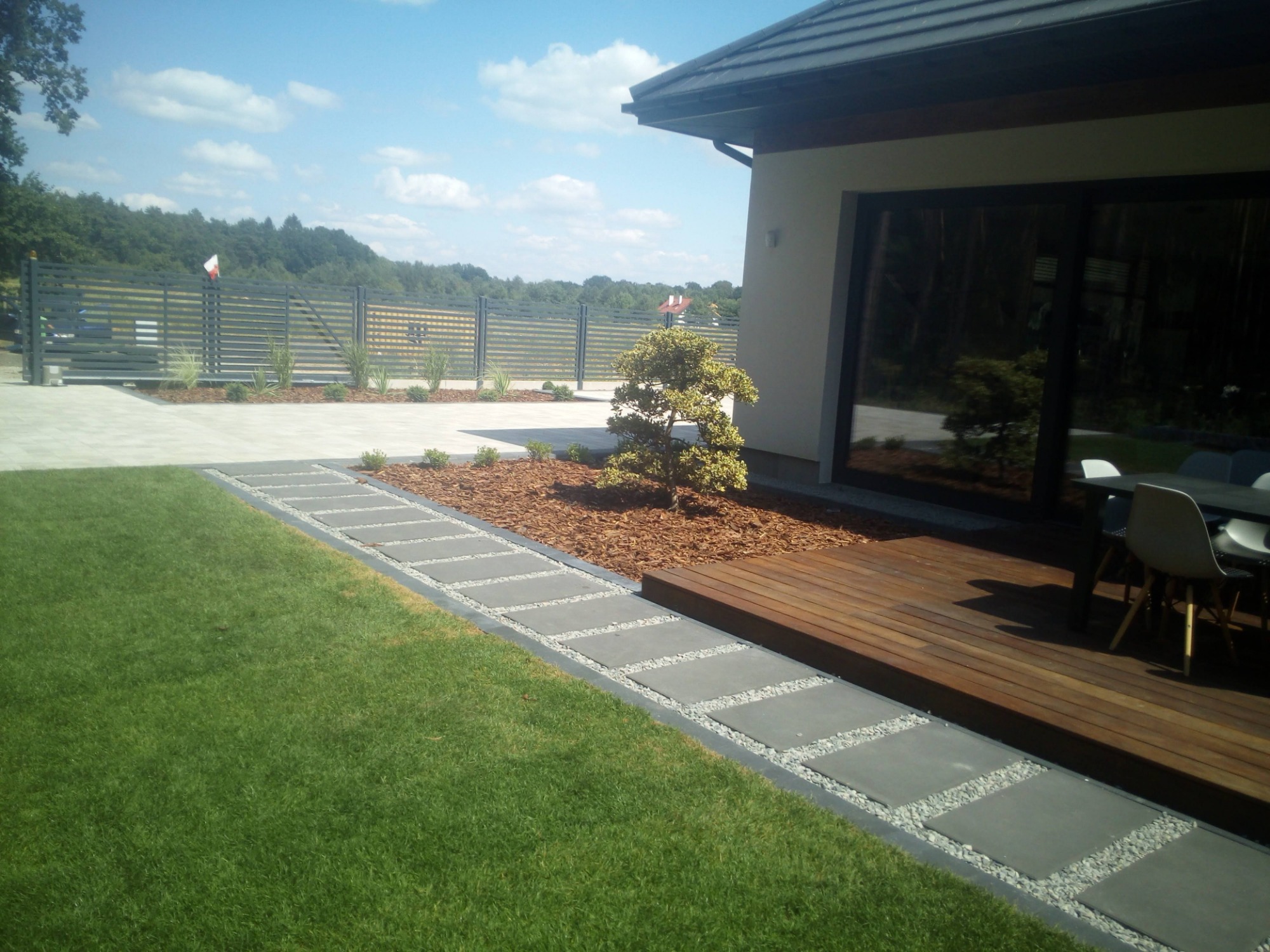 Nowoczesny ogród w Szczecinie z trawnikiem, ścieżką z płyt betonowych z wypełnieniem żwirowym, rabatą z korą i ozdobnym drzewkiem oraz tarasem drewnianym przylegającym do domu.