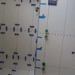 Białe płytki ścienne w trakcie układania, z niebieskimi krzyżykami dystansowymi i taśmą malarską, widoczne wystające przewody i otwory na instalacje.