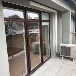 Biała jednostka zewnętrzna klimatyzacji Daikin zamontowana na balkonie z brązowymi drzwiami przesuwnymi i szarą balustradą.