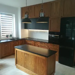 Kuchenna aranżacja z drewnianymi szafkami o wyraźnym usłojeniu, czarnymi blatami i wyspą na środku, oświetlona dwoma wiszącymi lampami w stylu industrialnym.