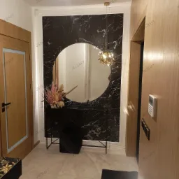 Nowoczesny przedpokój z czarnym marmurowym panelem, okrągłym lustrem i złotą lampą wiszącą, uzupełniony drewnianymi drzwiami i minimalistyczną konsolą z kwiatami.