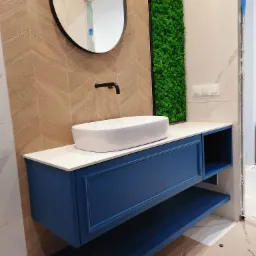 Niebieska szafka łazienkowa pod umywalkę nablatową, biały blat, lustro w czarnej ramie, ściana z drewnopodobnych płytek ułożonych w jodełkę, panel z zielonego mchu.