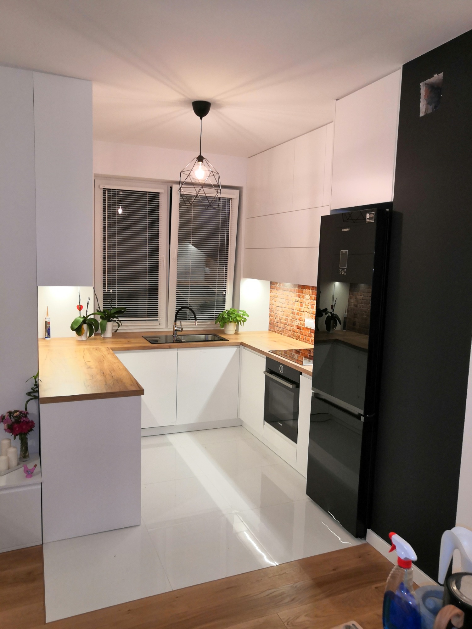 Nowoczesna kuchnia z białymi szafkami, drewnianym blatem, czarną lodówką i ceglaną ścianą nad blatem, oświetlona lampą wiszącą w stylu industrialnym.
