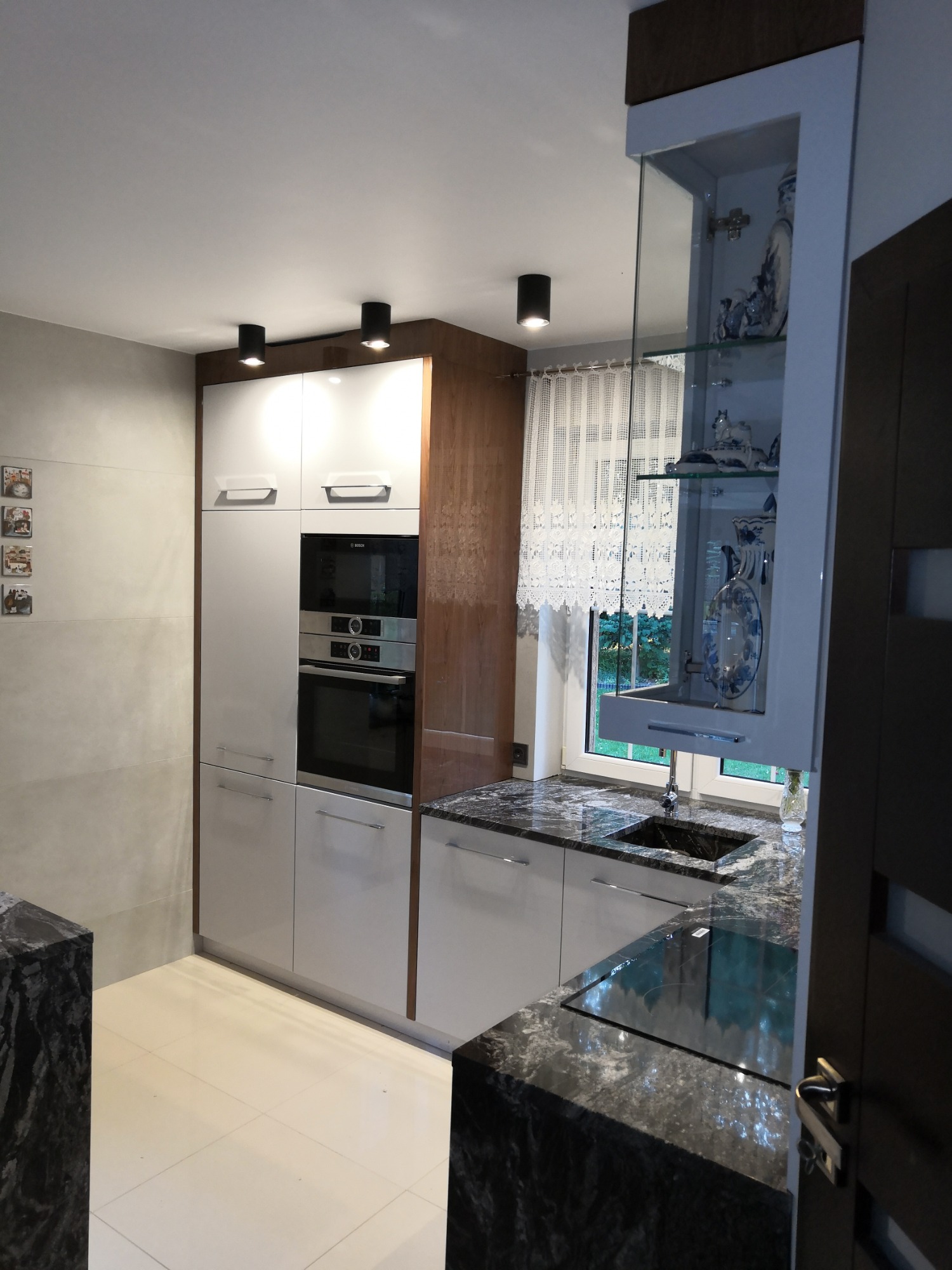 Biała kuchnia z zabudową piekarnika, szafką witrynową z porcelaną i blatem z ciemnego granitu, oświetlona czarnymi reflektorami sufitowymi, widok z perspektywy wejścia.