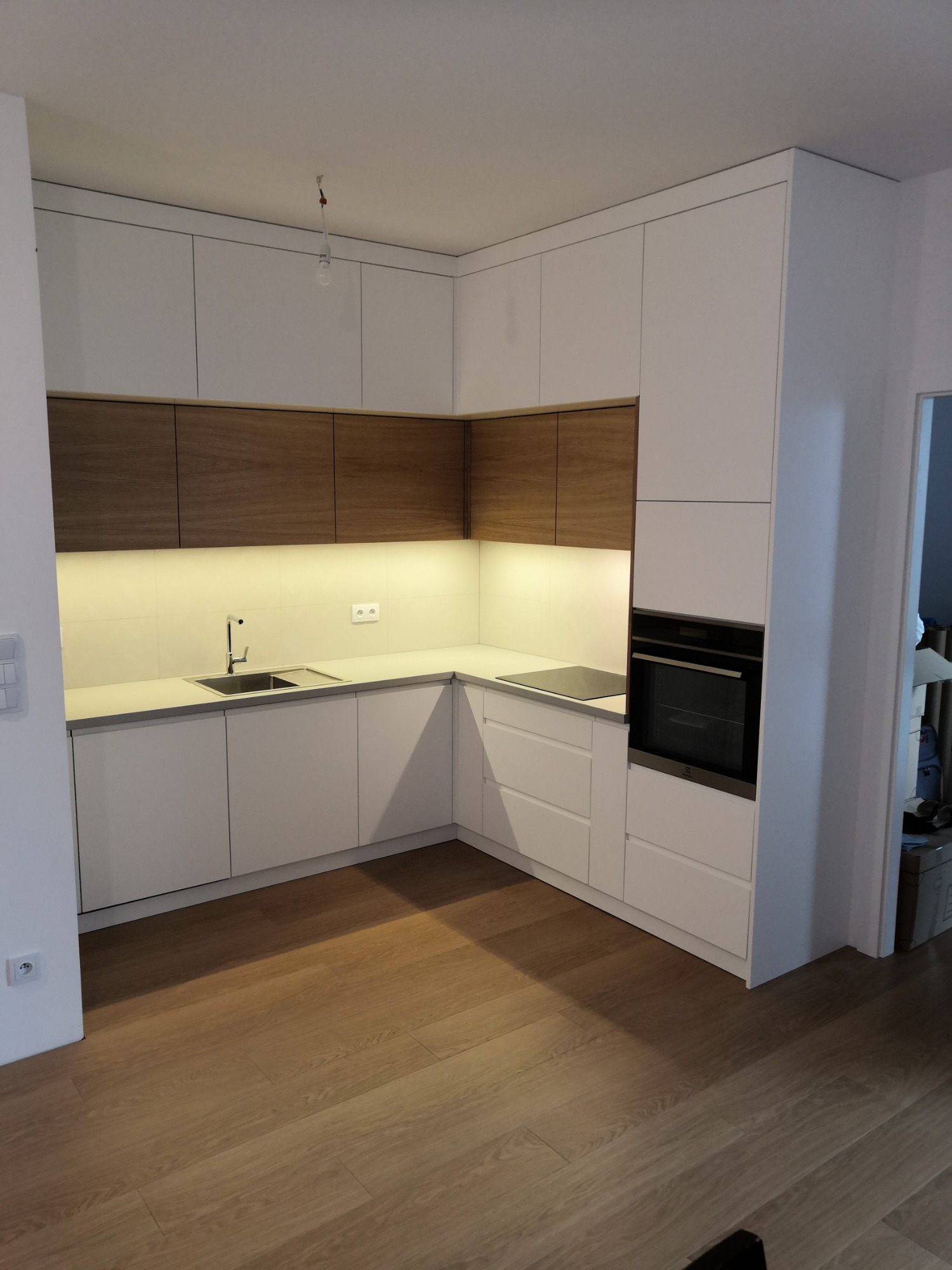 Nowoczesna kuchnia z białymi frontami i drewnianymi szafkami górnymi, oświetlenie LED pod szafkami, widoczny zlew, płyta indukcyjna i piekarnik, parkiet na podłodze, goła żarówka na suficie.