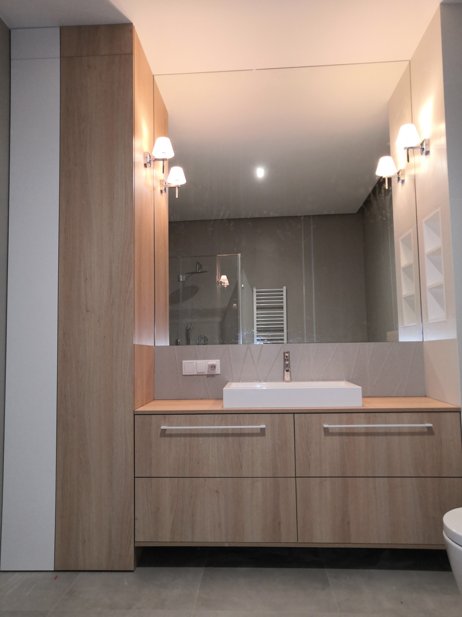 Nowoczesna łazienka z drewnianą zabudową meblową, prostokątną umywalką i dużym lustrem, w którym odbija się kabina prysznicowa i grzejnik drabinkowy.