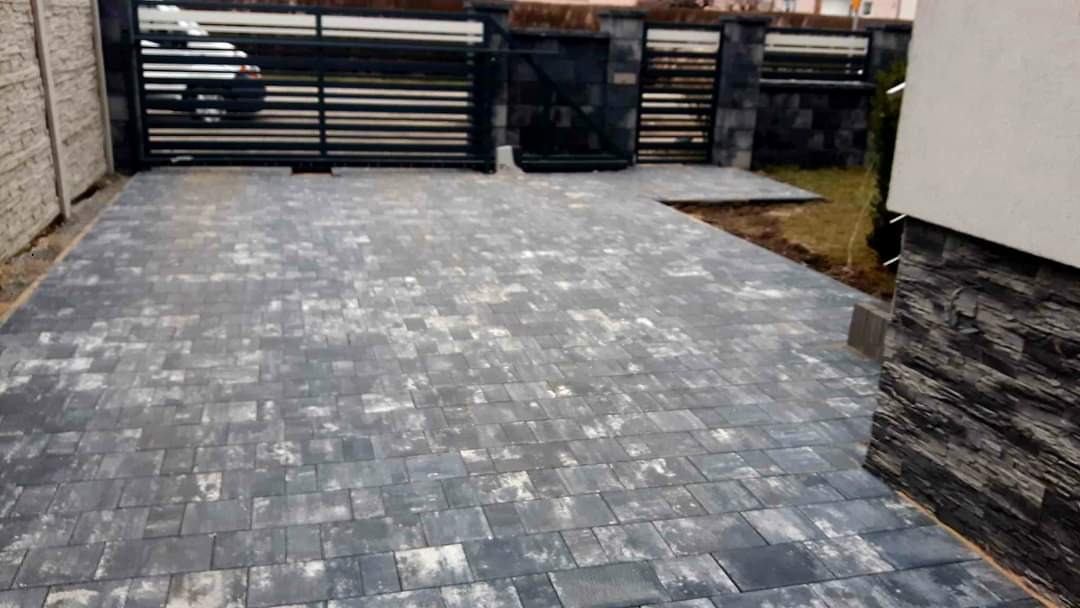 Ułożona szara kostka brukowa na podjeździe, widoczna brama wjazdowa z czarnymi, poziomymi przęsłami i fragment elewacji budynku z okładziną imitującą kamień.