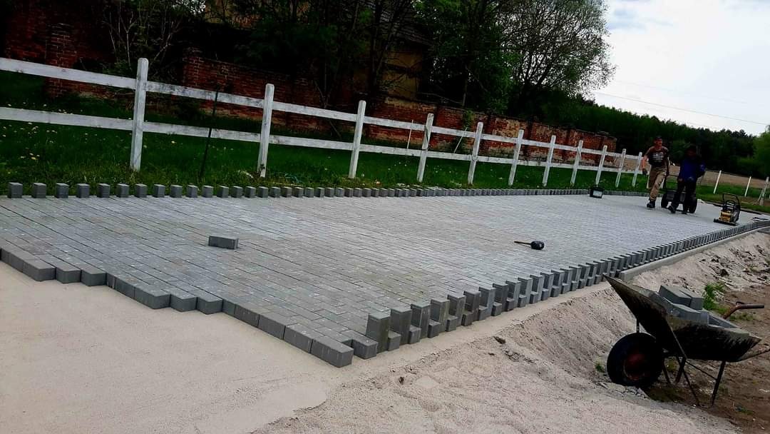 Układanie szarej kostki brukowej na podsypce piaskowej, widoczny fragment ułożonej powierzchni, obrzeża z wystającymi kostkami, w tle białe ogrodzenie i pracownicy z narzędziami.