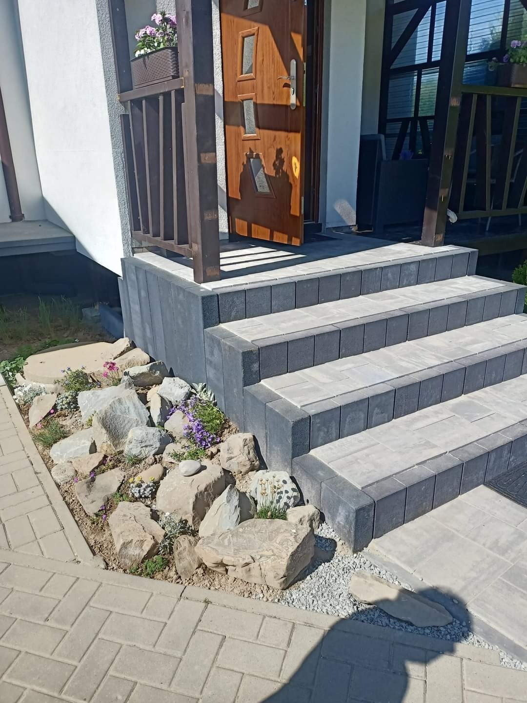 Schody wejściowe z kostki w odcieniach szarości, obok skalniak z kamieniami i roślinami, otwarte drzwi wejściowe z drewnianymi zdobieniami.