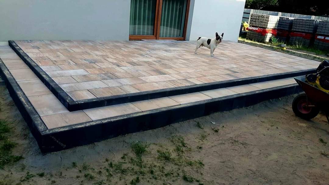 Wykończony taras z kostki brukowej w odcieniach beżu i szarości z czarnym obramowaniem, na którym stoi pies rasy buldog francuski, w tle okno i sterta kostki brukowej. Częściowo widoczna taczka.