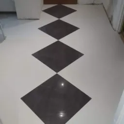 Biało-czarna podłoga z płytek ułożonych w romby w korytarzu, widoczna brązowa futryna drzwi i fragmenty ścian w stanie surowym.