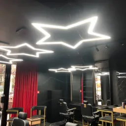 Wnętrze salonu fryzjerskiego z czarnymi ścianami i sufitem, oświetlone lampami LED w kształcie gwiazd, z widocznymi fotelami fryzjerskimi i schodami opartymi o okno.