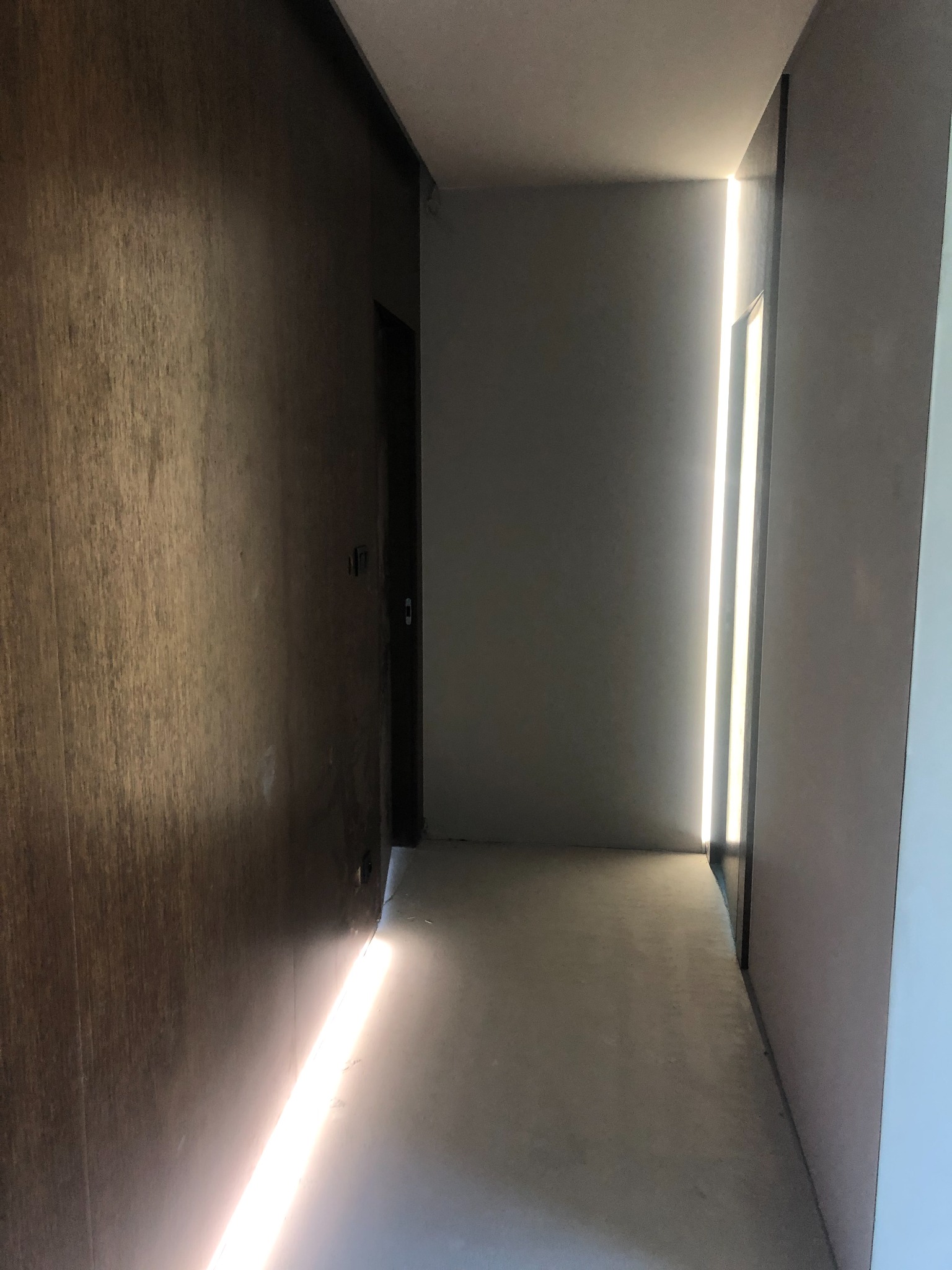 Korytarz w nowoczesnym domu jednorodzinnym z podświetleniem LED wzdłuż ściany, tworzącym pionową linię światła. Widoczna drewniana okładzina ściany po lewej stronie i surowa podłoga.