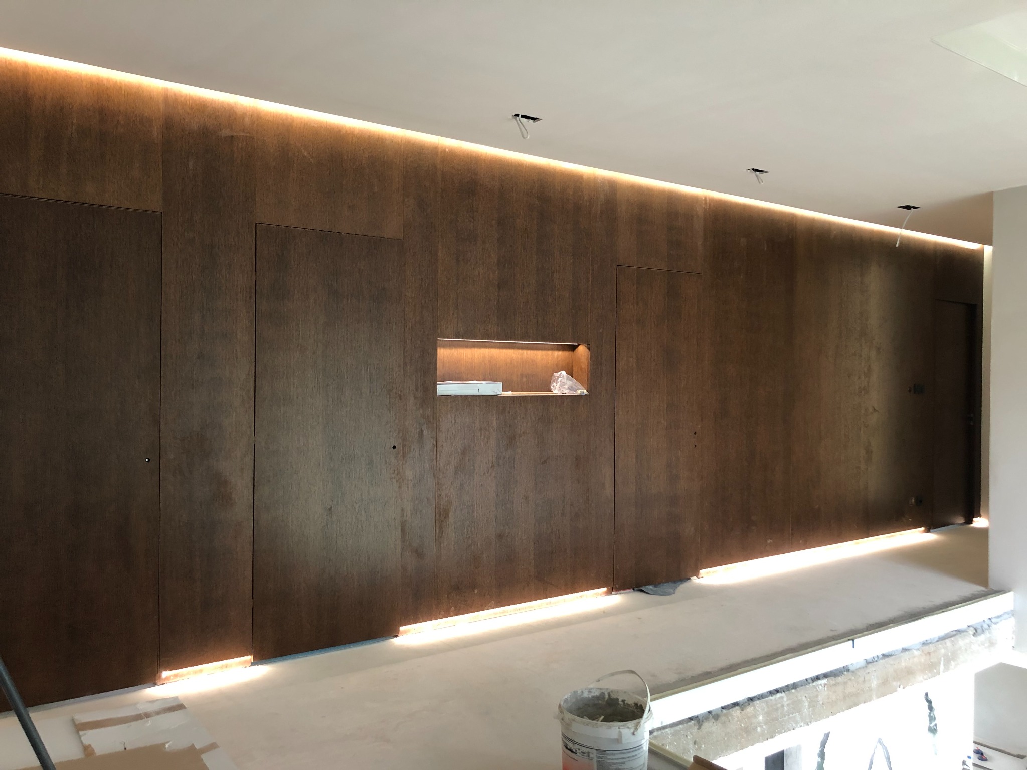 Korytarz w trakcie wykańczania z ciemnobrązową, drewnianą ścianą z ukrytymi drzwiami i wnęką z podświetleniem LED, widoczne przewody elektryczne wystające z sufitu, na podłodze wiadro z zaprawą.
