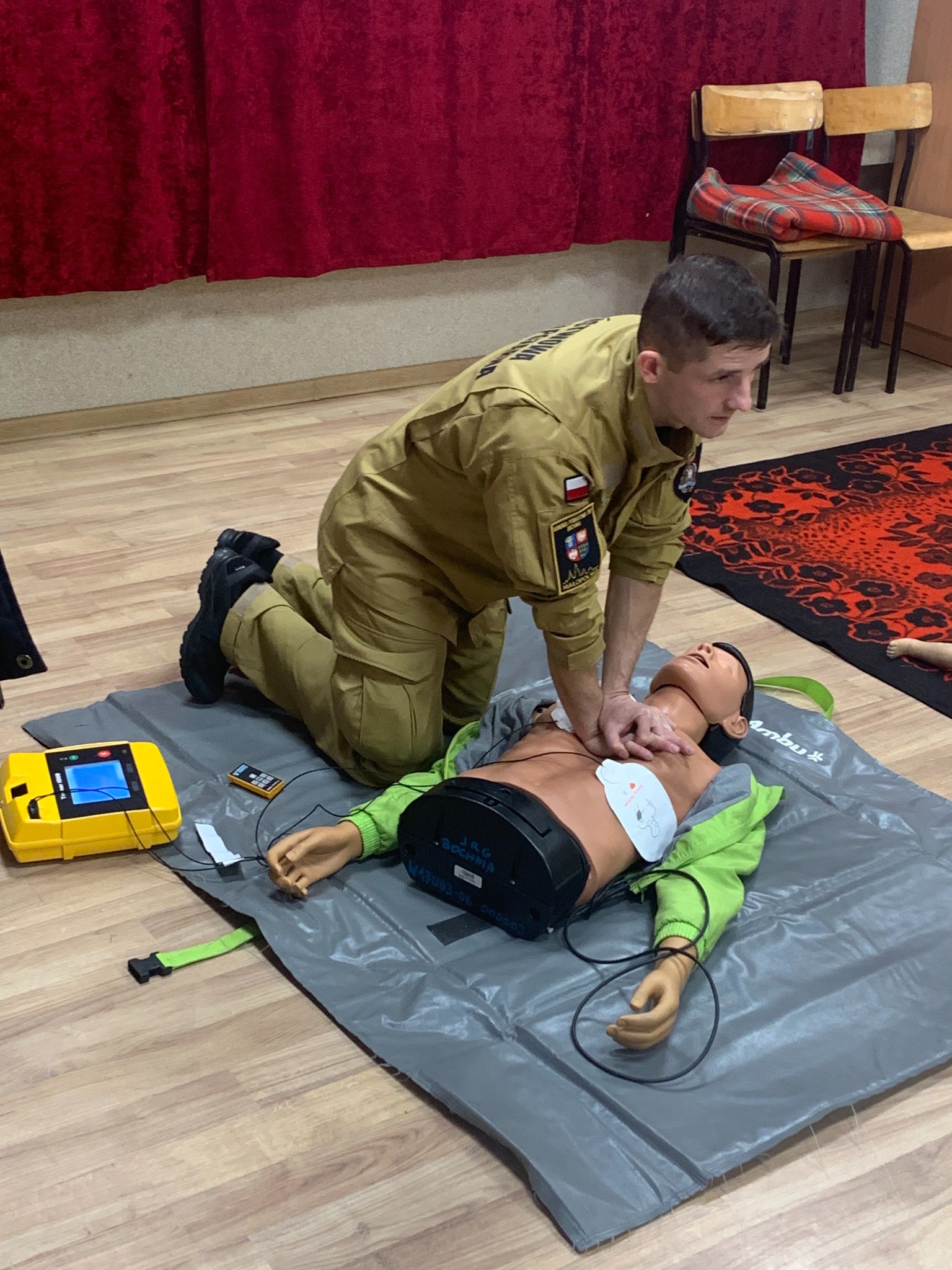 Instruktor w mundurze demonstruje resuscytację krążeniowo-oddechową na manekinie z podłączonym defibrylatorem. Ćwiczenia z pierwszej pomocy w pomieszczeniu.