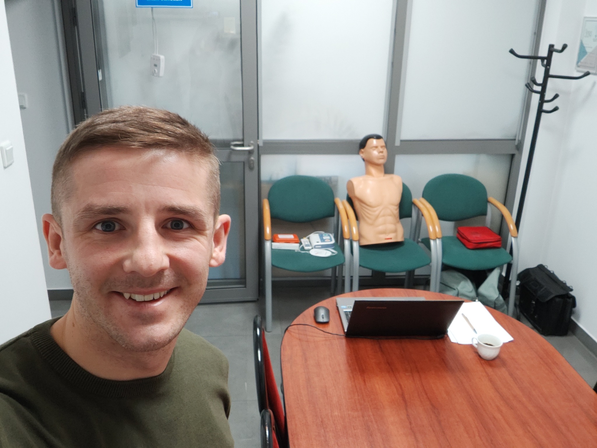 Uśmiechnięty mężczyzna na tle sali szkoleniowej z manekinem do ćwiczeń RKO, laptopem i kubkiem kawy na stole. Widoczne krzesła i defibrylator.