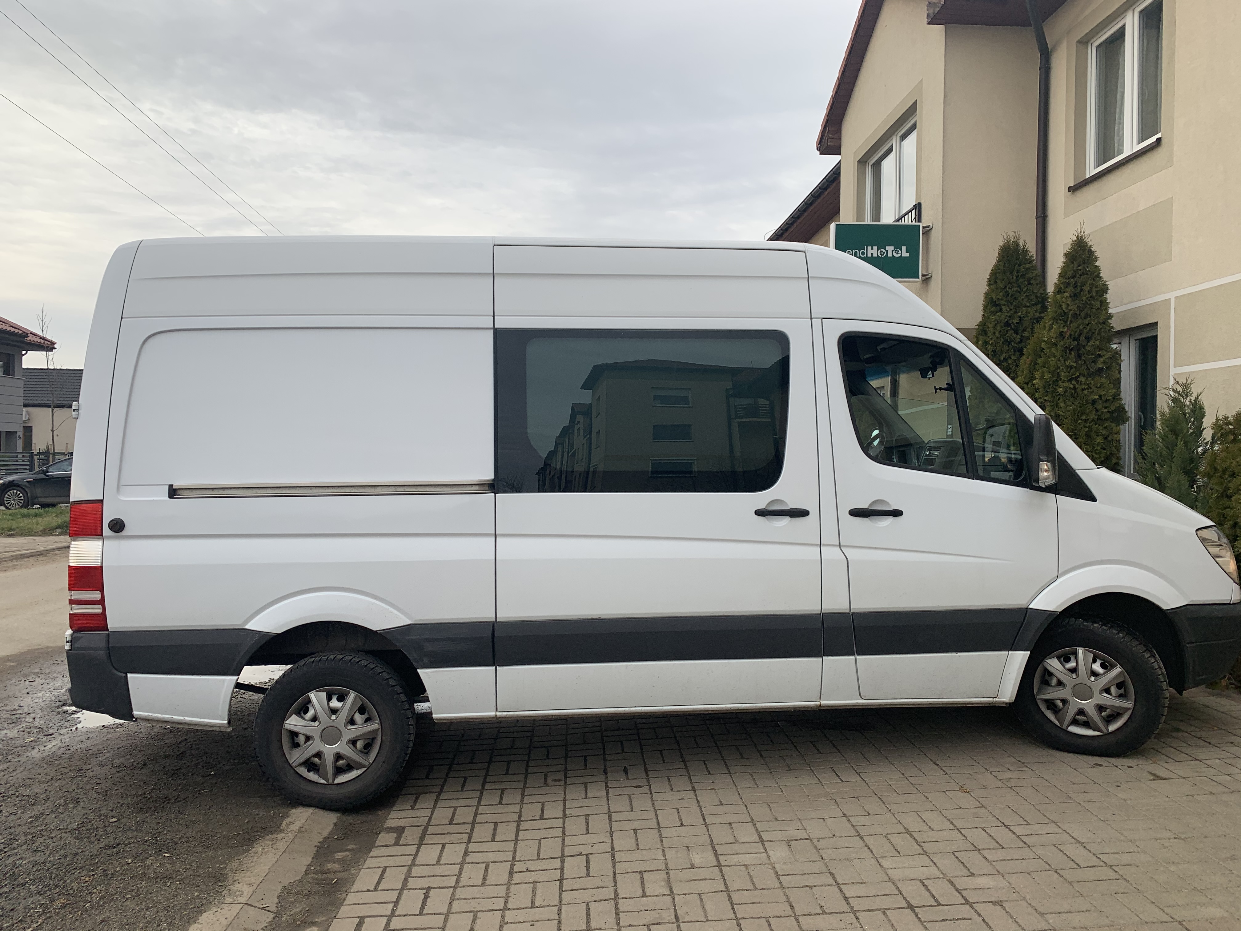 Biały bus dostawczy zaparkowany na chodniku z kostki brukowej, z widocznym znakiem hotelu w tle.