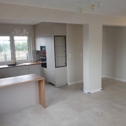 Usługi budowlane TOMAX Tomasz Cieśla - Jasne wnętrze po remoncie: widok na kuchnię z wyspą, świeżo pomalowane ściany i listwy przypodłogowe zabezpieczone taśmą malarską, drzwi wewnętrzne w tle.